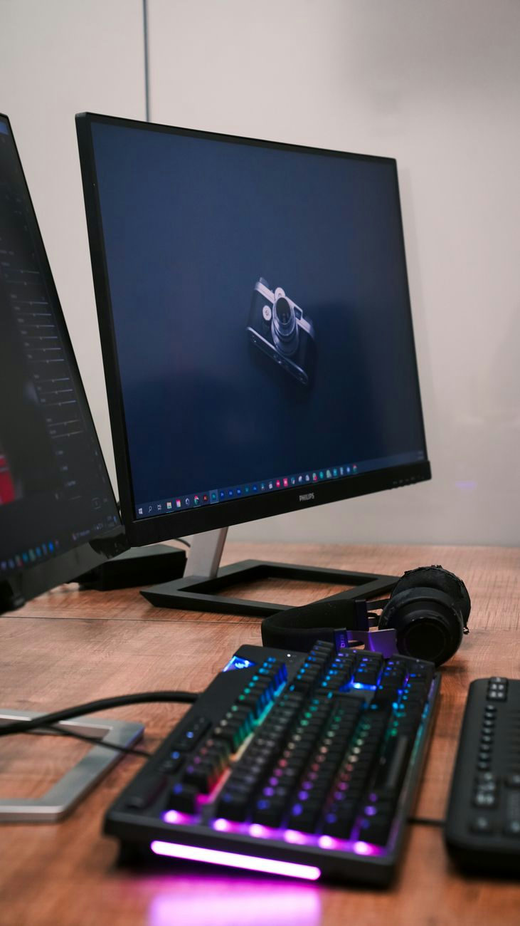 A sleek desktop setup featuring two monitors, an RGB-lit keyboard, and headphones on a wooden desk.