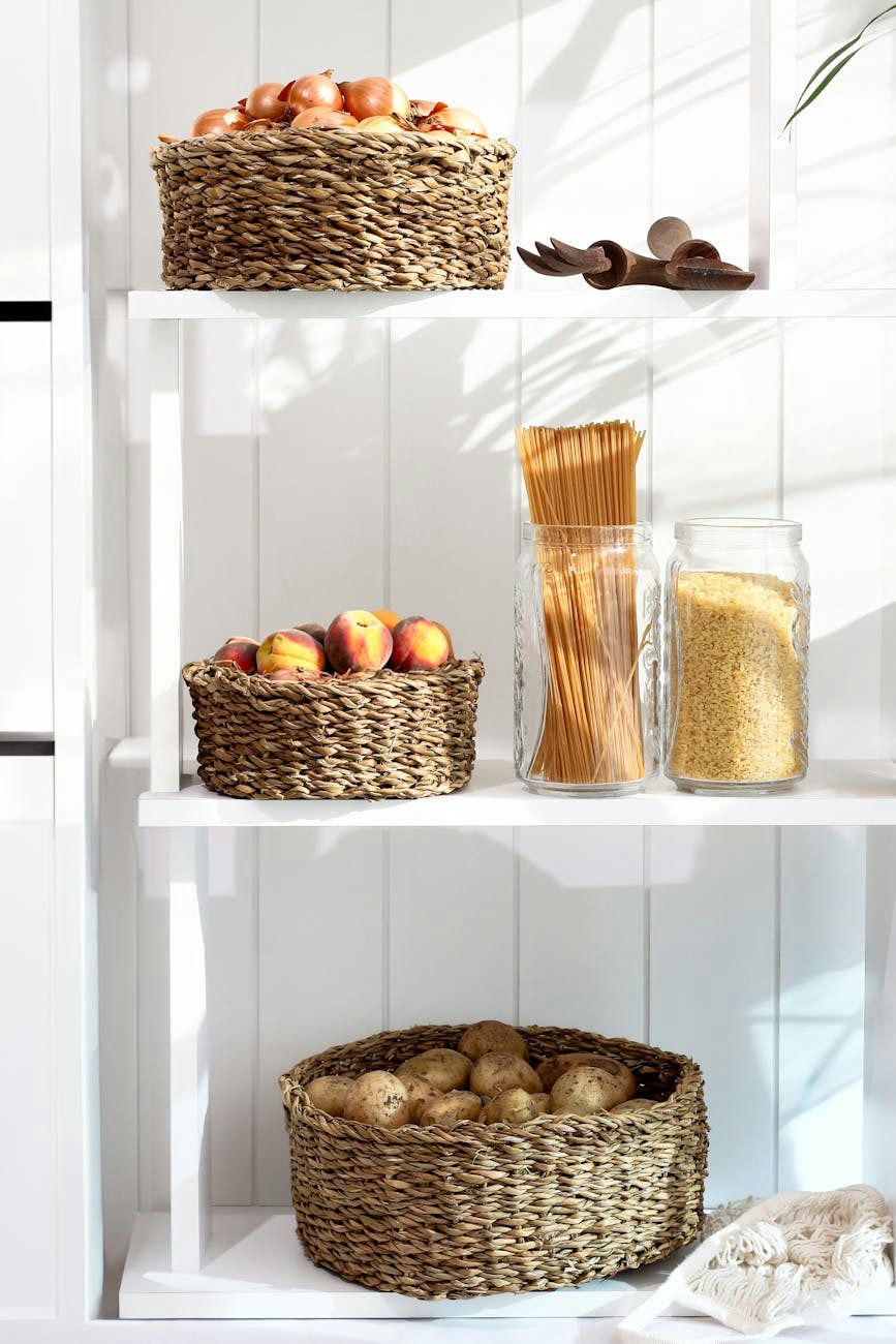 A neatly organized pantry shelf with wicker baskets holding apples, peaches, and potatoes, alongside pasta and grains.