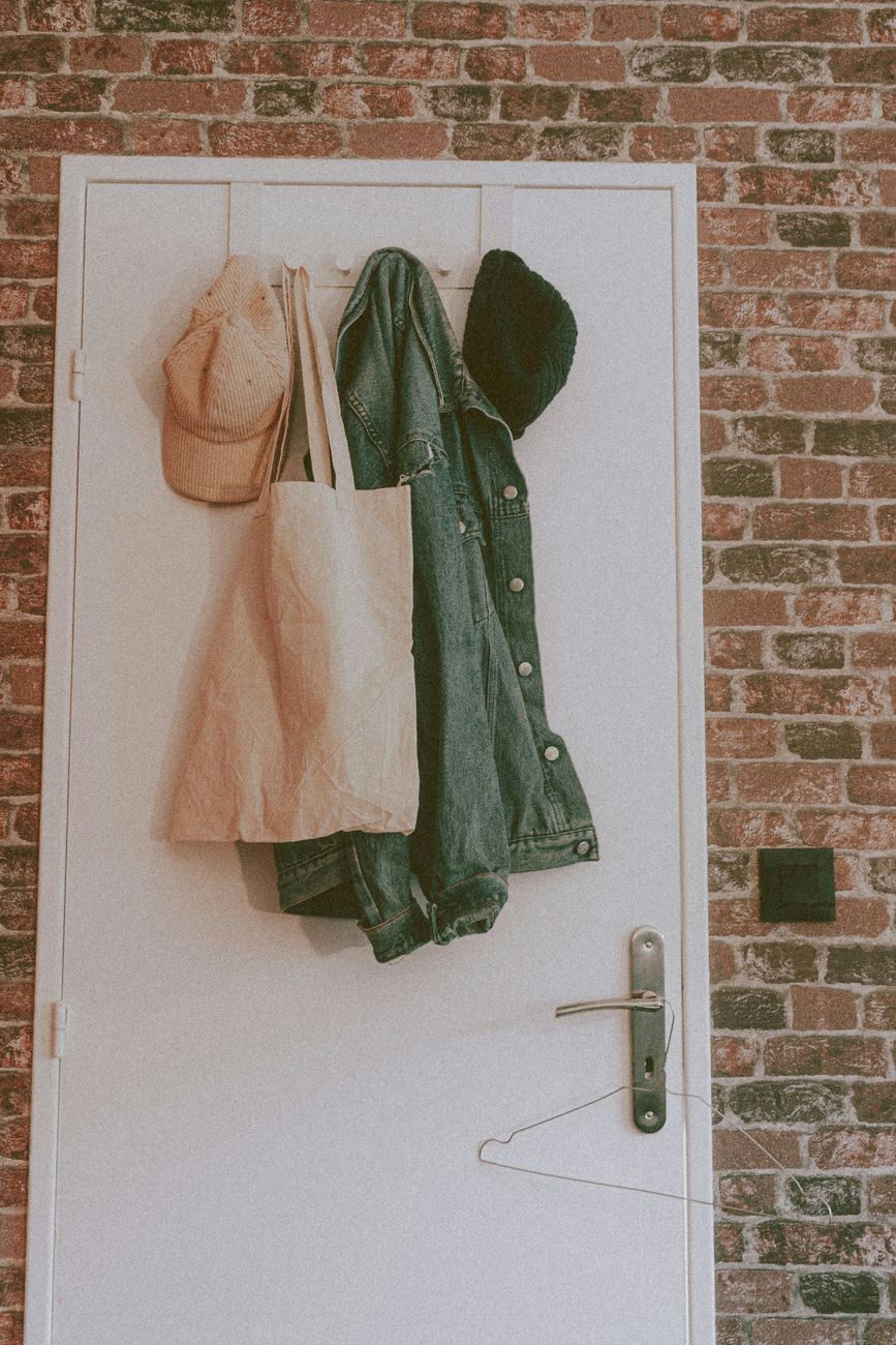 Light pink bag and cap with denim coat and warm hat on concrete hanger on white door in room with bricks walls