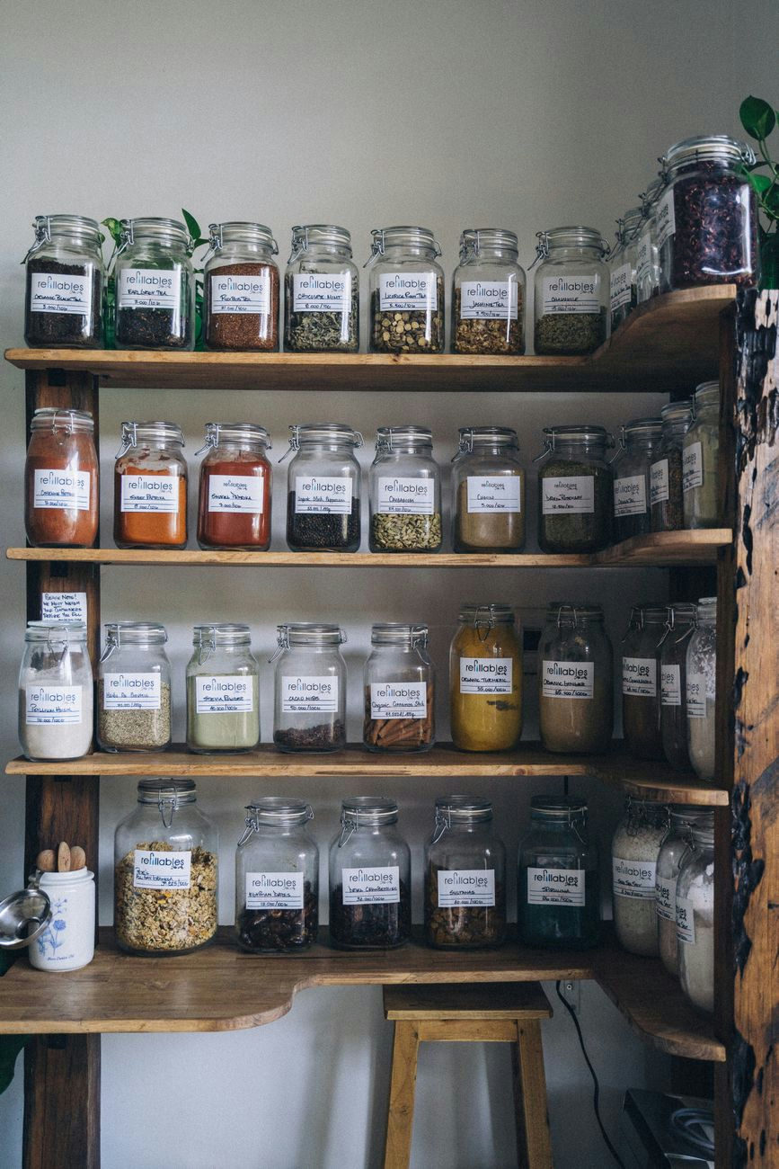 A wooden shelf displaying assorted spices in clear refillable glass jars, encouraging eco-friendly storage.