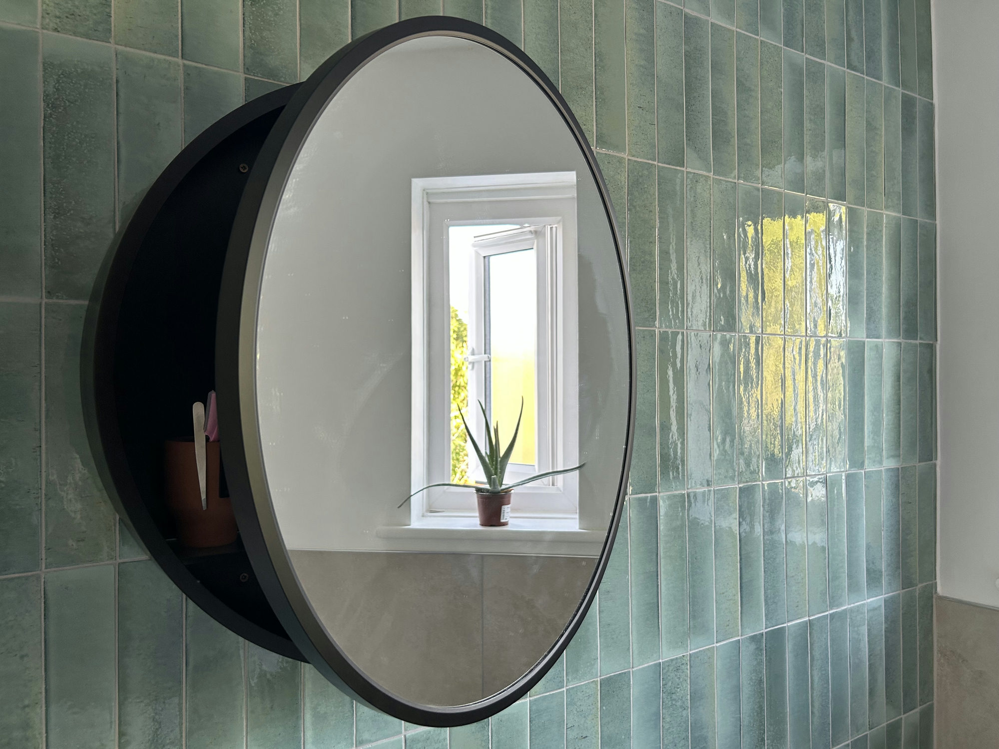 A circular mirrored bathroom cabinet with a plant in the window shown in the reflection.