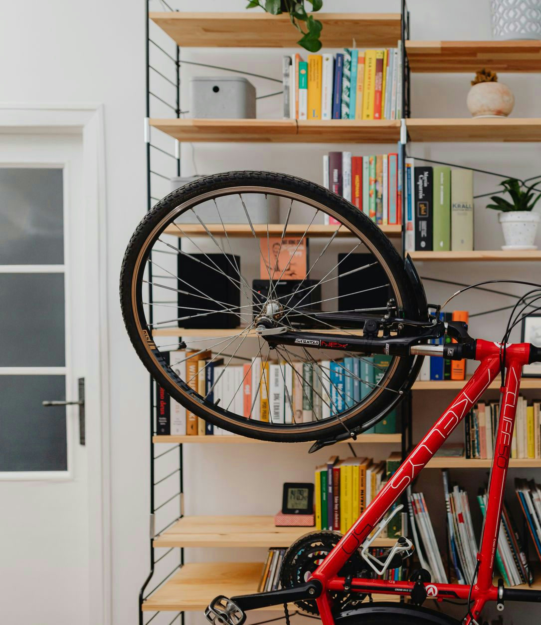 A man in casual attire positions a red bicycle indoors near a bookshelf filled with books.